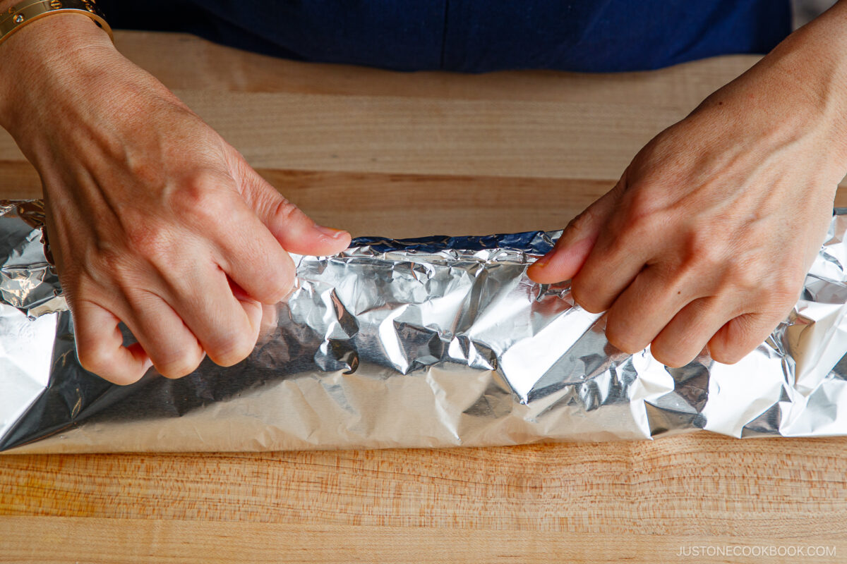 A person’s hands tightly wrapping a long item in aluminum foil on a wooden surface. The foil is being pressed and folded to secure the contents.