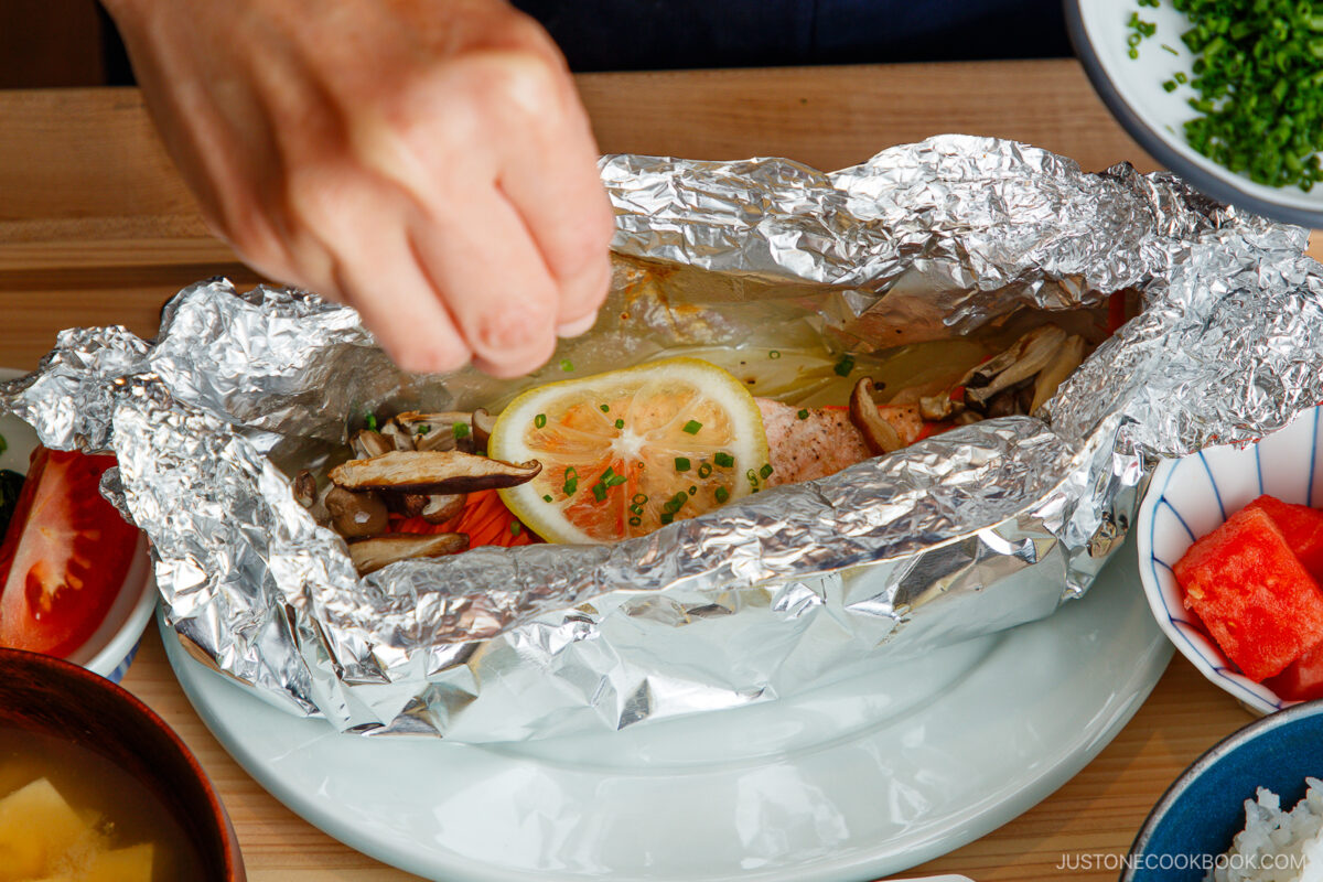 A hand opens a foil packet containing cooked fish topped with lemon slices, mushrooms, and chopped chives, surrounded by plates of soup, sliced tomatoes, and other side dishes on a wooden table.