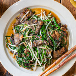 A plate of stir-fried beef liver with bean sprouts and green vegetables, served with chopsticks on a wooden table. A bowl of rice and small dish of pickles are nearby.