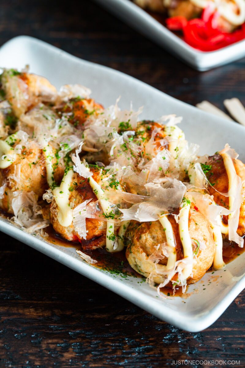 A rectangular plate of takoyaki, Japanese octopus balls, topped with mayonnaise, takoyaki sauce, dried bonito flakes, and sprinkled green herbs, on a dark wooden table.