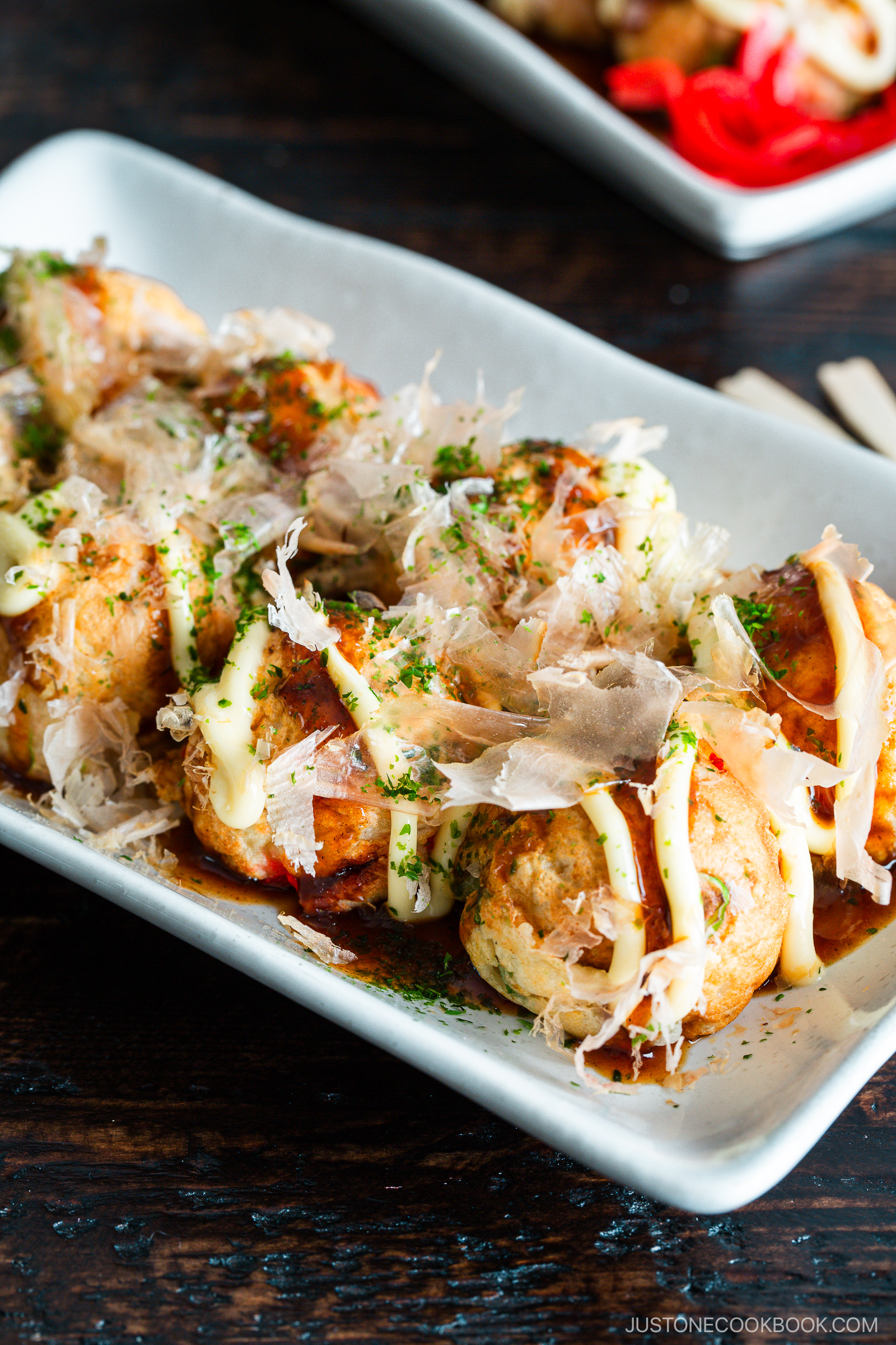 A rectangular plate of takoyaki, Japanese octopus balls, topped with mayonnaise, takoyaki sauce, dried bonito flakes, and sprinkled green herbs, on a dark wooden table.