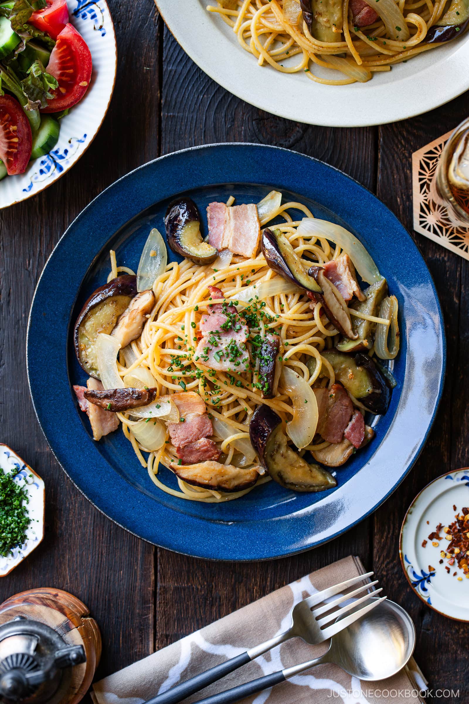 A blue plate of spaghetti topped with slices of bacon, sautéed eggplant, and onions, garnished with chopped herbs, sits on a dark wooden table next to a fork, spoon, napkin, and a side salad.
