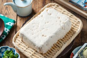 A block of fresh tofu rests on a woven bamboo tray, surrounded by various small dishes and utensils on a wooden table.