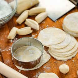 A wooden board with round dumpling wrappers, dough balls, a rolling pin, a metal cutter, cut dough pieces, and a bowl of flour, showing the process of making dumpling wrappers.