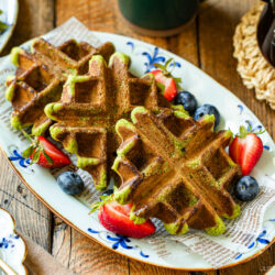 A plate of matcha waffles is served with halved strawberries and blueberries on a rustic wooden table. The waffles are arranged on decorative paper, with coffee and more waffles visible in the background.