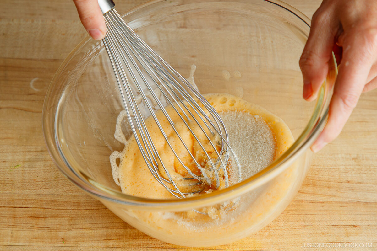 A person whisking sugar into beaten eggs in a glass bowl on a wooden surface.