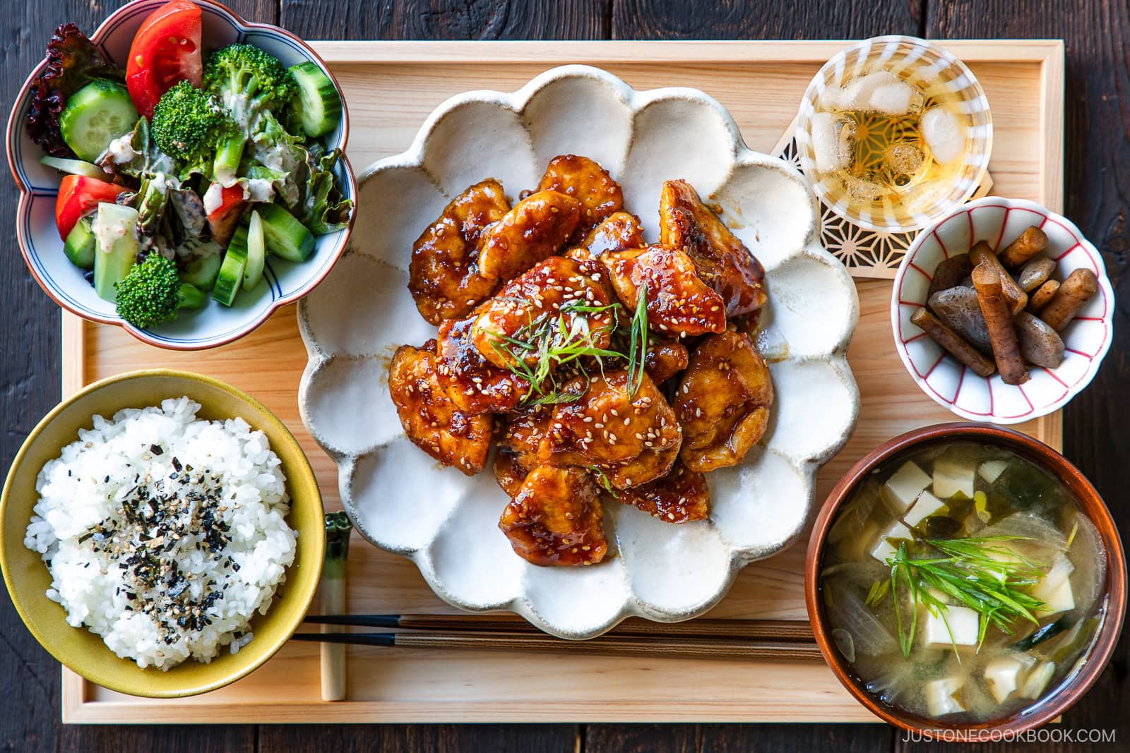 A Japanese meal set on a wooden tray with glazed chicken, steamed rice, miso soup, mixed salad, and a small bowl of pickled vegetables, all neatly arranged in separate dishes.