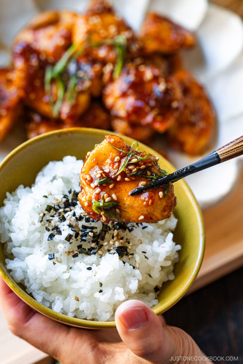A hand holds a bowl of white rice topped with sesame seeds and nori flakes, while chopsticks lift a piece of glazed chicken garnished with green onions and sesame seeds. Blurred plate of more chicken in the background.