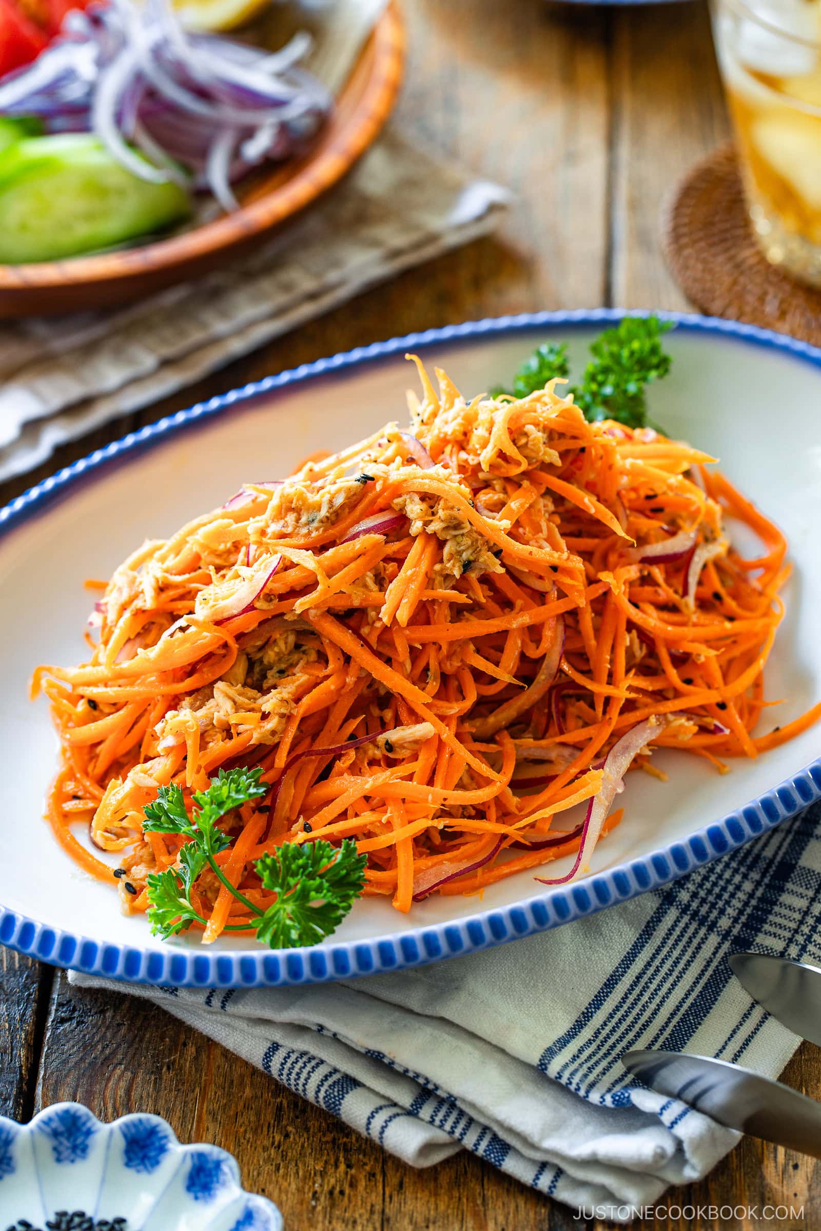 A plate of shredded carrot salad mixed with chicken, garnished with parsley, sits on a wooden table. In the background, there is a plate with sliced red onions, cucumber, and tomato.