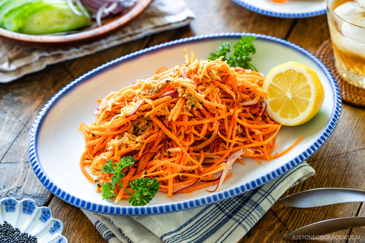 A white oval plate with shredded carrot salad garnished with parsley and half a lemon, set on a striped napkin. Other dishes and a glass of drink are partly visible in the background.