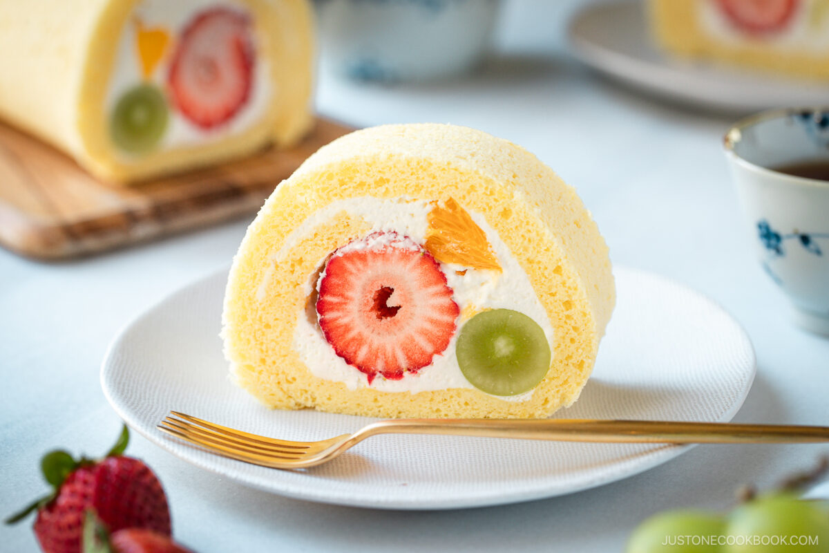 A slice of fluffy yellow Swiss roll cake filled with whipped cream, a strawberry, a green grape, and a piece of orange sits on a white plate with a gold fork. Whole strawberries and another cake slice are in the background.