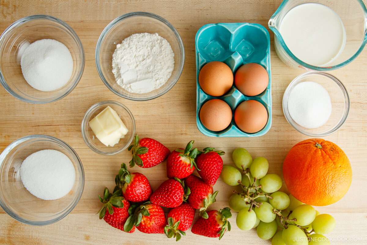 A top-down view of baking ingredients on a wooden surface, including bowls of sugar, flour, butter, eggs in a carton, milk in a jug, strawberries, green grapes, and an orange.