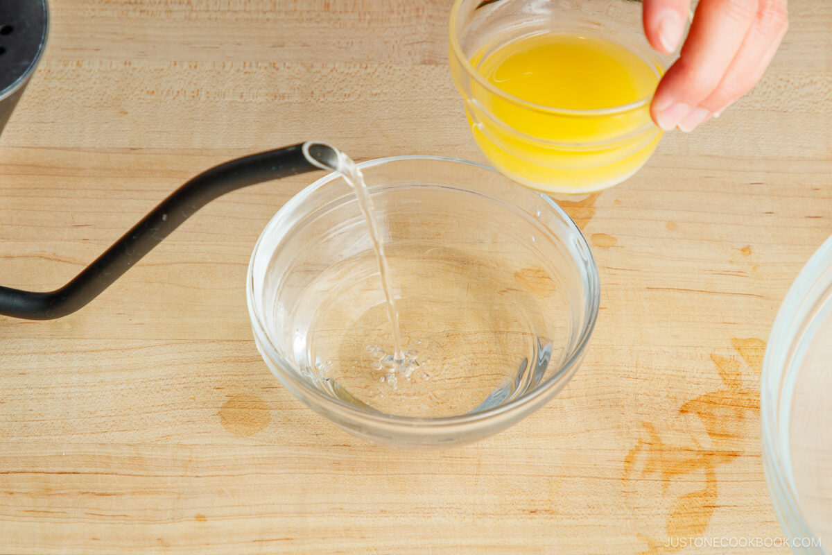A hand holds a small bowl of melted butter above a glass bowl, while hot water is being poured into the glass bowl on a wooden surface.