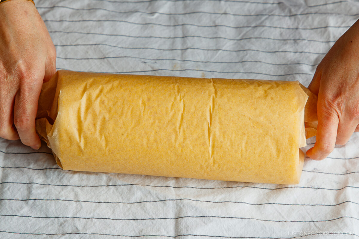 Two hands holding a rolled-up log of parchment paper containing dough, placed on a white cloth with thin black stripes.