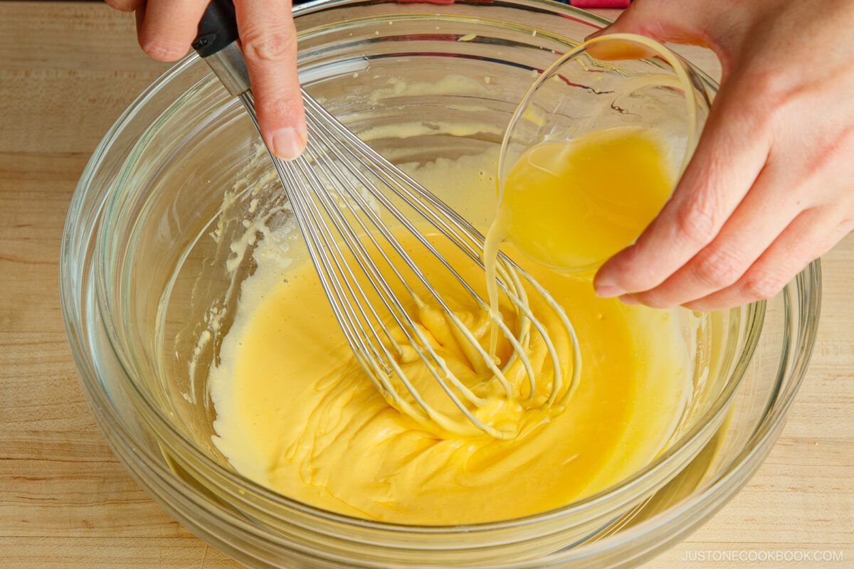 A person whisks a yellow batter in a glass bowl while pouring a cup of melted butter into the mixture on a wooden surface.
