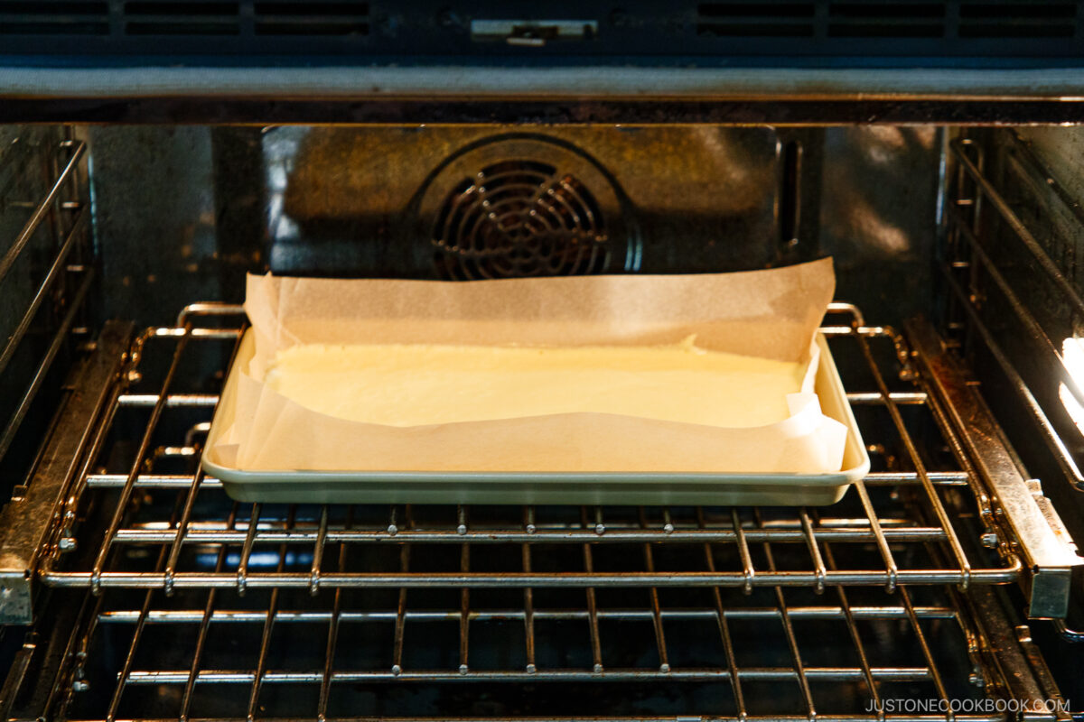 A rectangular baking pan lined with parchment paper and filled with batter is placed on the middle rack of an oven, baking.