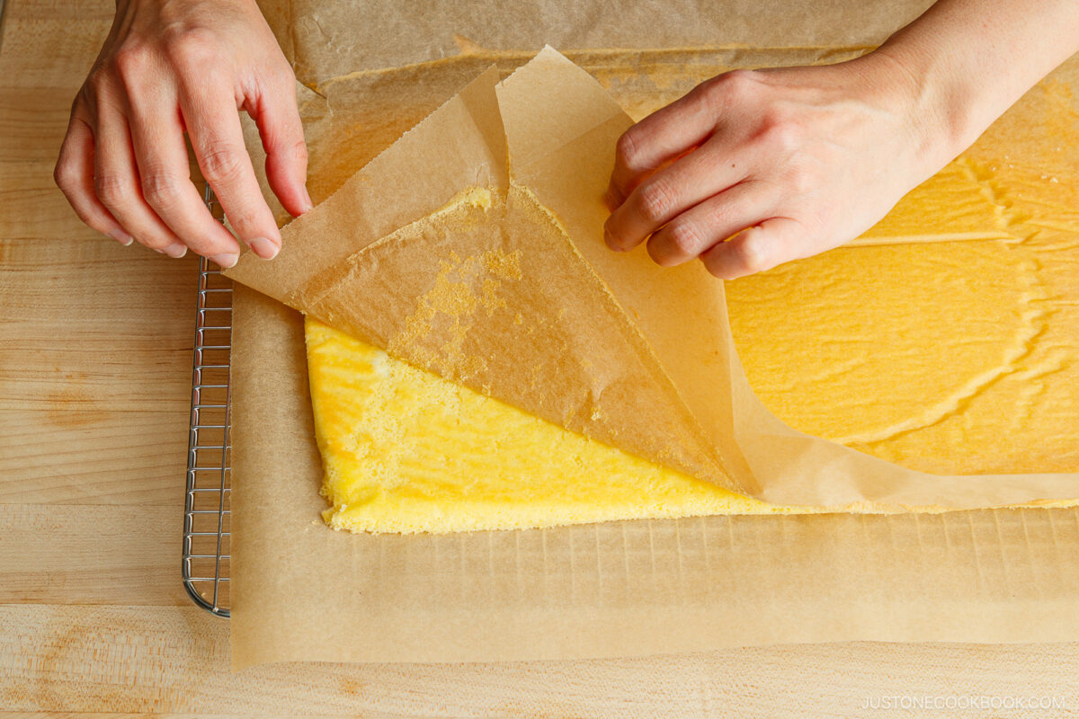 Two hands peel back a sheet of parchment paper from a thin, rectangular layer of yellow sponge cake on a cooling rack over a wooden surface.
