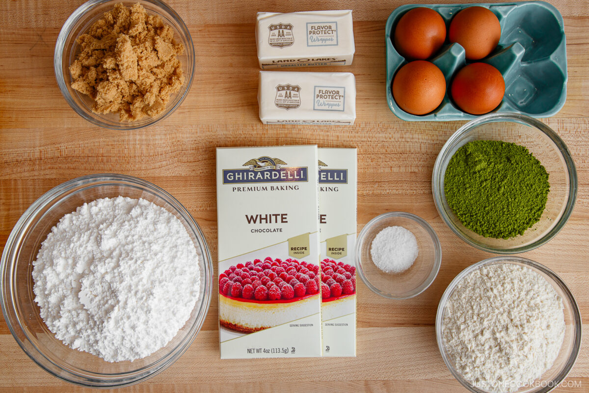 Ingredients for baking laid out on a wooden surface: brown sugar, butter sticks, eggs, matcha powder, white chocolate bars, flour, powdered sugar, and a small bowl of salt.