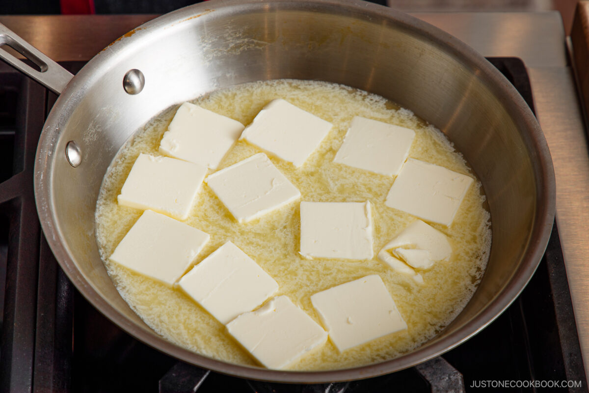 Sliced pieces of butter melting in a stainless steel pan on a stovetop.