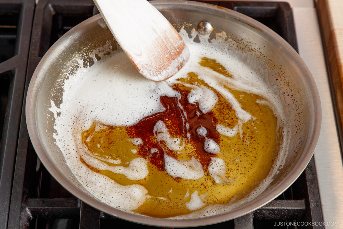 A stainless steel pan on a stovetop contains melted browned butter, with light foam and brown specks. A wooden spatula rests in the pan, stirring the mixture.