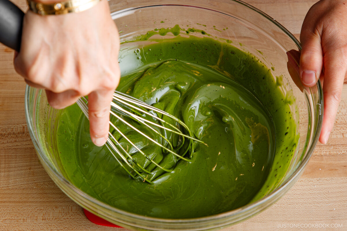Close-up of hands whisking a thick, vibrant green matcha mixture in a clear glass bowl on a wooden surface.