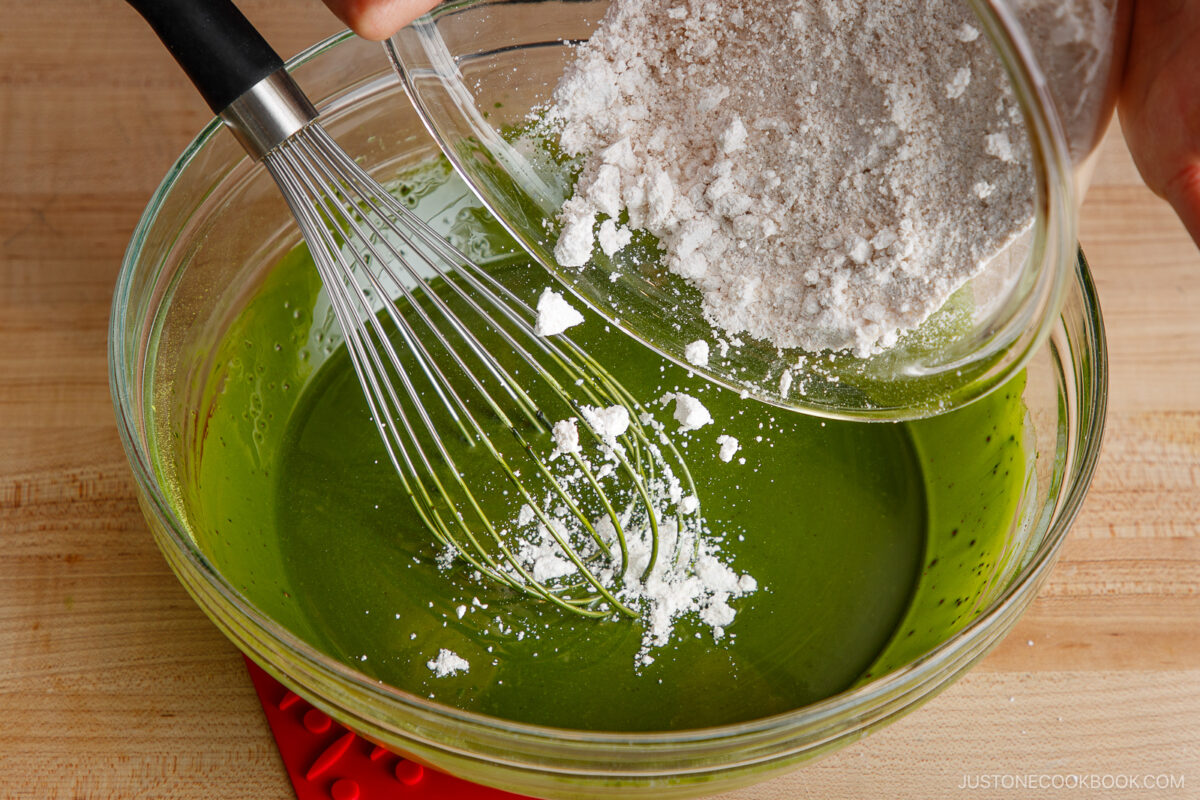 A hand pours a bowl of flour into a large glass bowl filled with green matcha mixture while a whisk rests inside, preparing batter on a wooden surface.