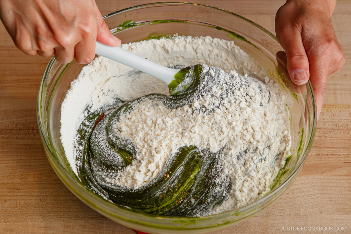 A person mixes flour into a green matcha batter using a white spatula in a clear glass bowl on a wooden surface.