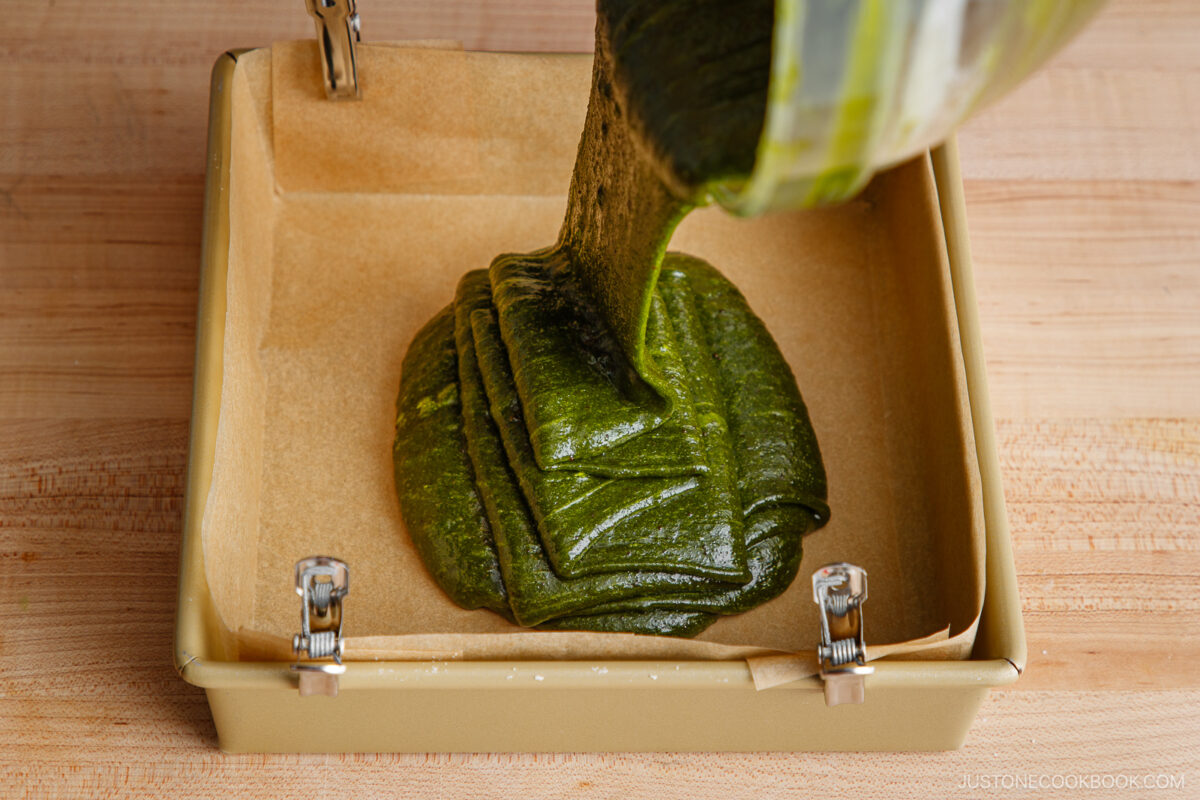 A thick, green batter is being poured from a bowl into a square, parchment-lined baking pan secured with metal clips, on a wooden surface.