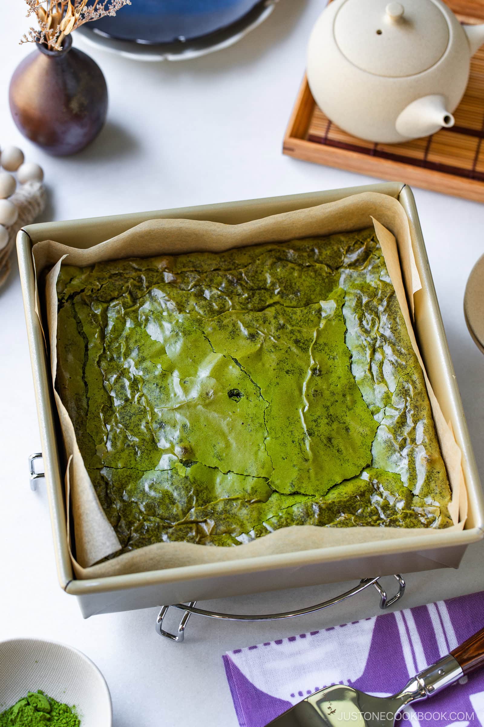 A square matcha green tea cake in a parchment-lined baking pan sits on a table with a teapot, teacup, plate, and a small bowl of matcha powder nearby. The cake has a shiny, cracked surface.