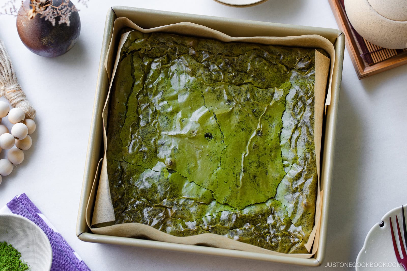 A square baking pan lined with parchment paper contains a baked sheet of shiny, green matcha brownies. Surrounding the pan are bowls of matcha powder and white beans, and a wooden utensil.