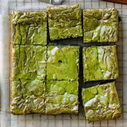 A batch of matcha brownies sliced into nine squares sits on parchment paper, with a small dish of green matcha powder and a purple-patterned napkin nearby.