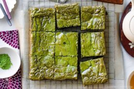 A batch of matcha brownies sliced into nine squares sits on parchment paper, with a small dish of green matcha powder and a purple-patterned napkin nearby.