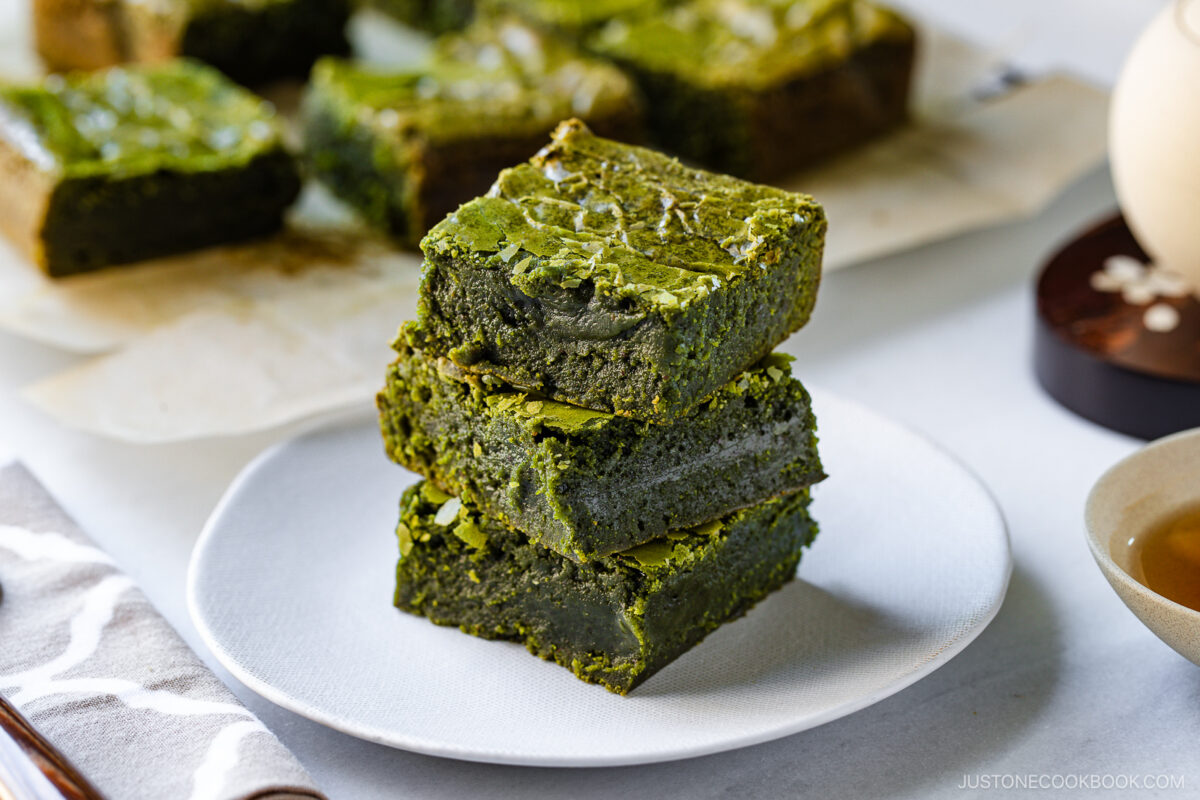 Three thick, green matcha brownies are stacked on a white plate, with more brownies and a cup of tea in the background. The brownies have a rich, moist texture and a marbled surface.