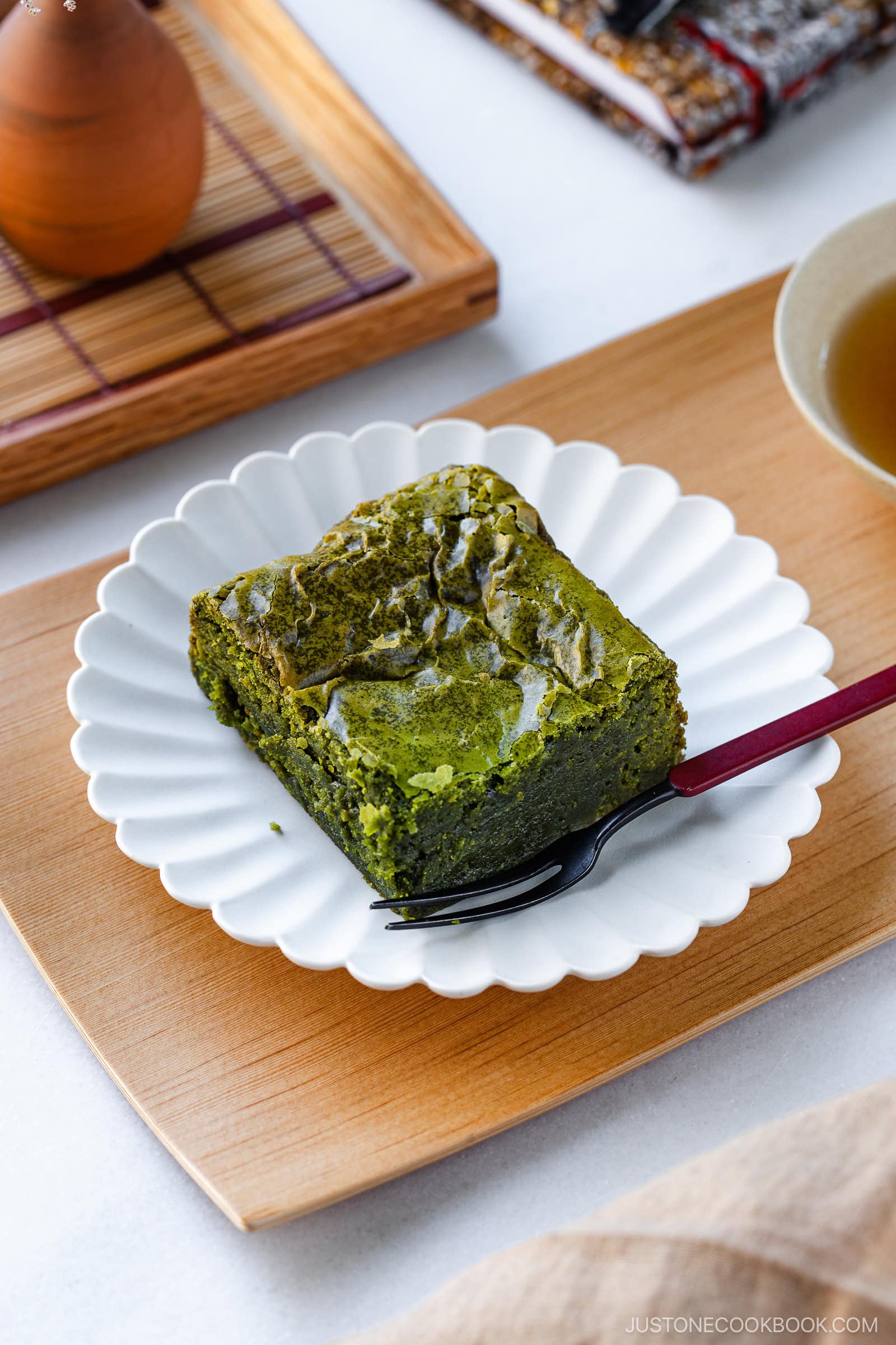 A piece of matcha green tea brownie sits on a white scalloped plate with a black fork, on a wooden tray. A cup of tea and a brown teapot are partially visible in the background.