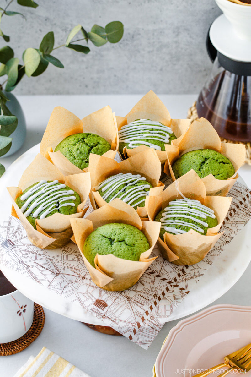 Eight green muffins, some drizzled with white icing, are arranged on a round marble stand with parchment wrappers. Greenery and coffee pots are visible in the background, creating a cozy setting.