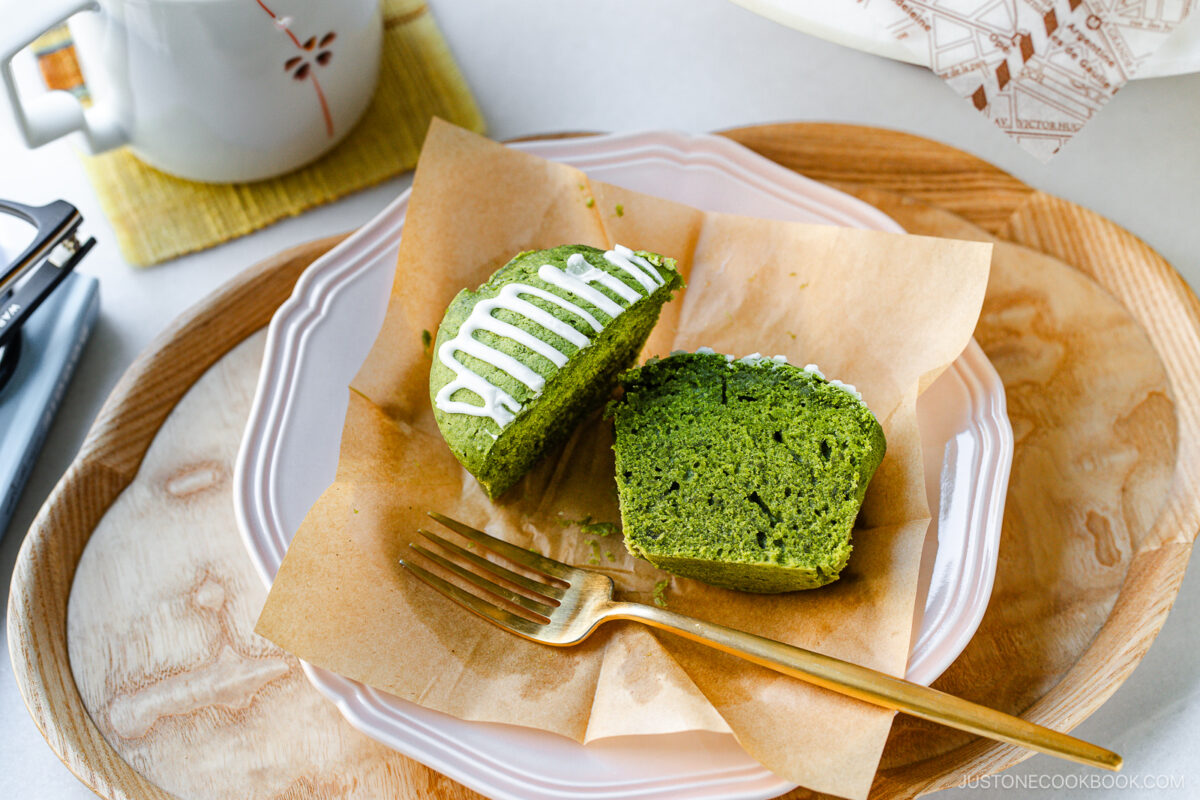 A green matcha muffin with white icing is cut in half on parchment paper, placed on a pink plate with a gold fork. A teapot and a wrapped pastry are on the wooden tray beside it.