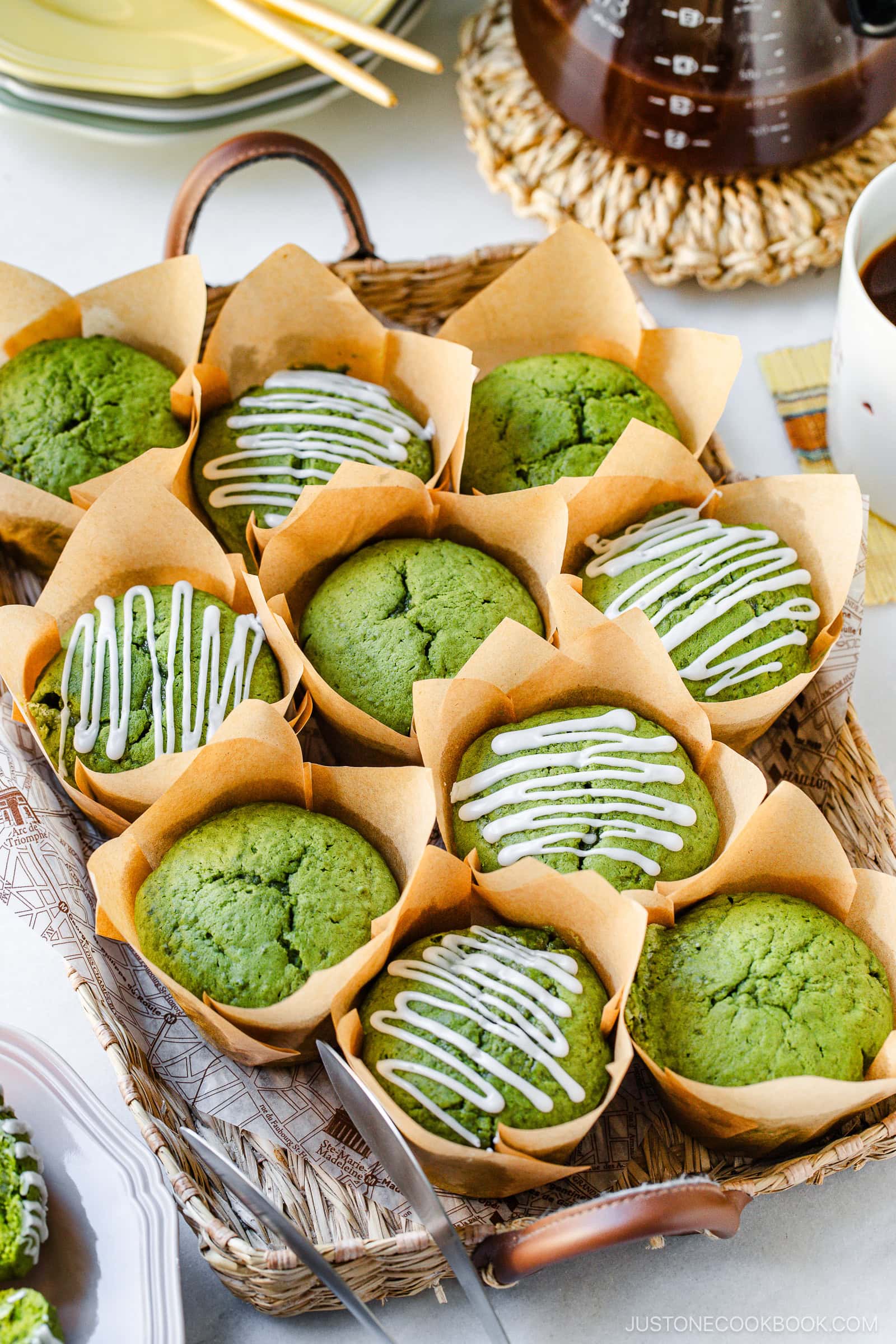 A basket of matcha muffins in brown parchment wrappers, some topped with white icing drizzle and others plain, arranged neatly on a table next to plates and a pot of coffee.