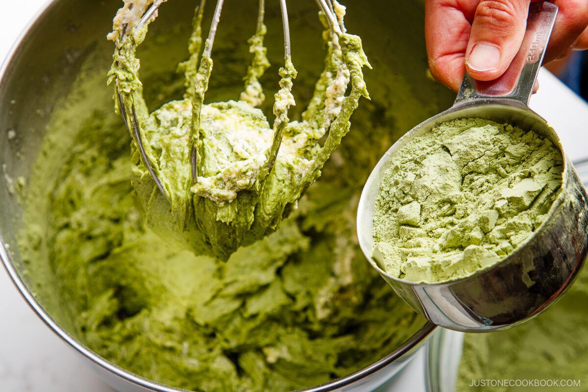 A hand holds a measuring cup filled with green matcha powder above a mixing bowl with batter and a whisk attachment coated in the same green mixture.