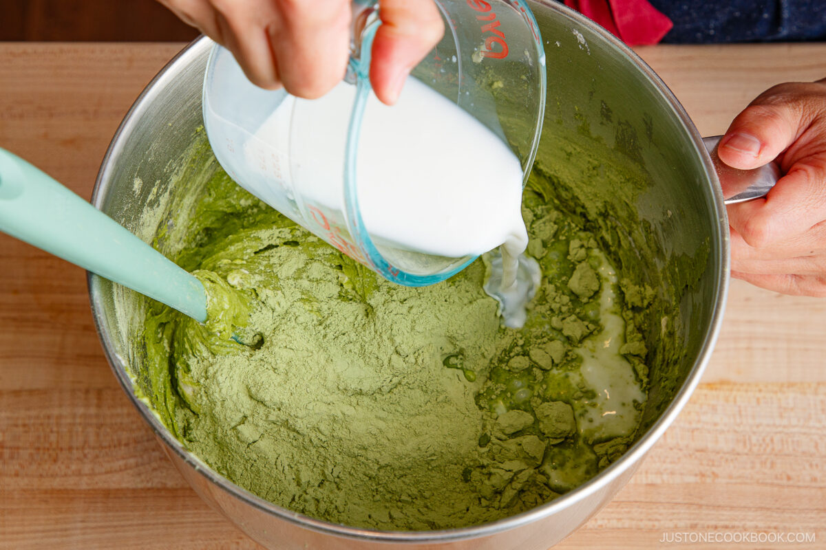 A person pours milk from a measuring cup into a mixing bowl filled with green matcha powder, preparing a batter with a green spatula on a wooden surface.