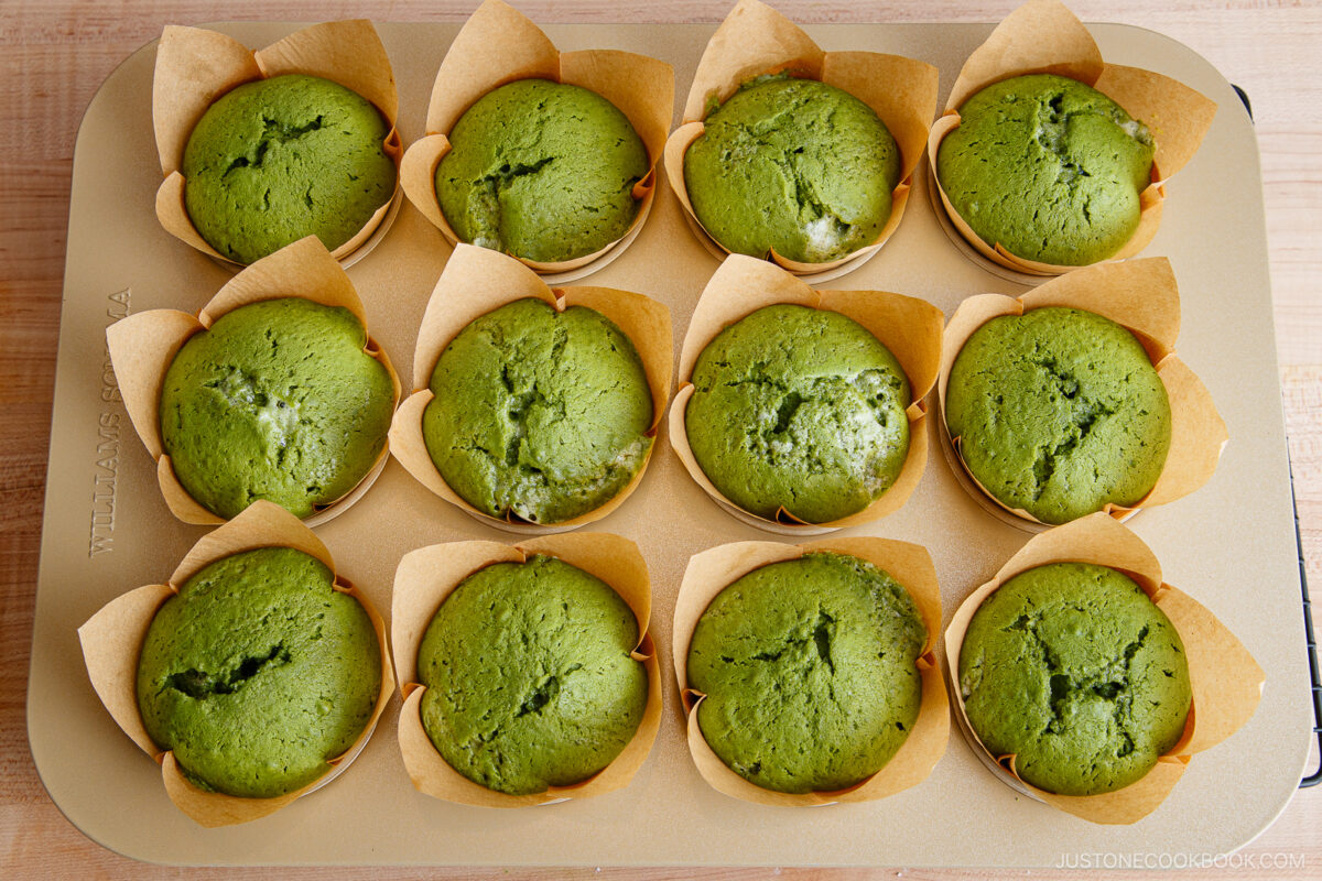 A baking tray holds twelve freshly baked green muffins in brown paper liners, arranged neatly in three rows of four. The muffins have slightly cracked tops and a soft texture.
