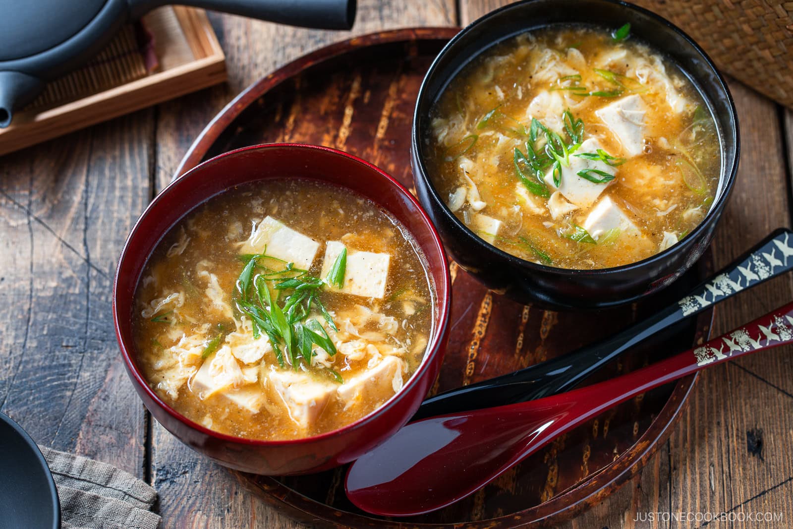 Two bowls of miso soup with tofu, egg, and green onions sit on a wooden tray. Red and black lacquered spoons rest beside the bowls, showcasing a cozy, traditional Japanese meal on a rustic table.