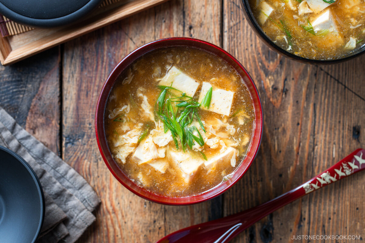 A bowl of egg drop soup with tofu cubes and sliced green onions, served in a red bowl on a rustic wooden table, with a red spoon and other dishes nearby.