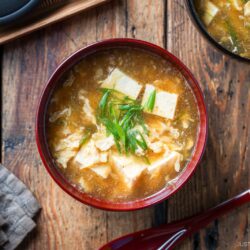 A bowl of egg drop soup with tofu cubes and sliced green onions, served in a red bowl on a rustic wooden table, with a red spoon and other dishes nearby.