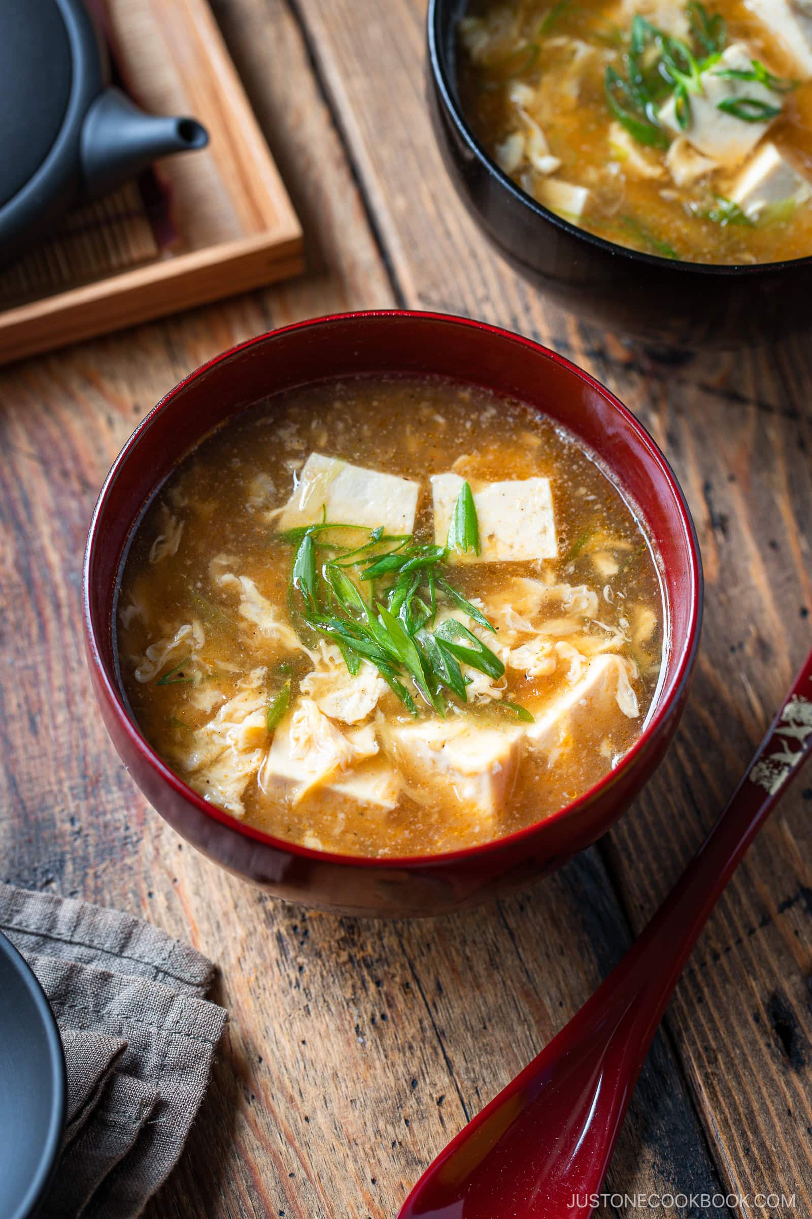 A bowl of hot and sour soup with tofu, egg ribbons, and green onions, served in a red bowl on a wooden table with a red spoon and another bowl of soup in the background.