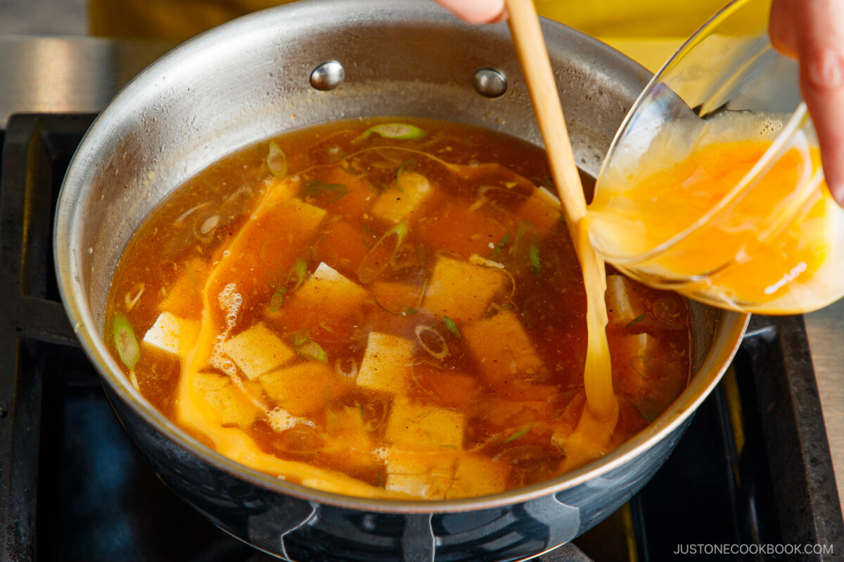 A hand pours beaten eggs from a bowl into a pan of simmering soup with tofu cubes, green onions, and broth on a stovetop.