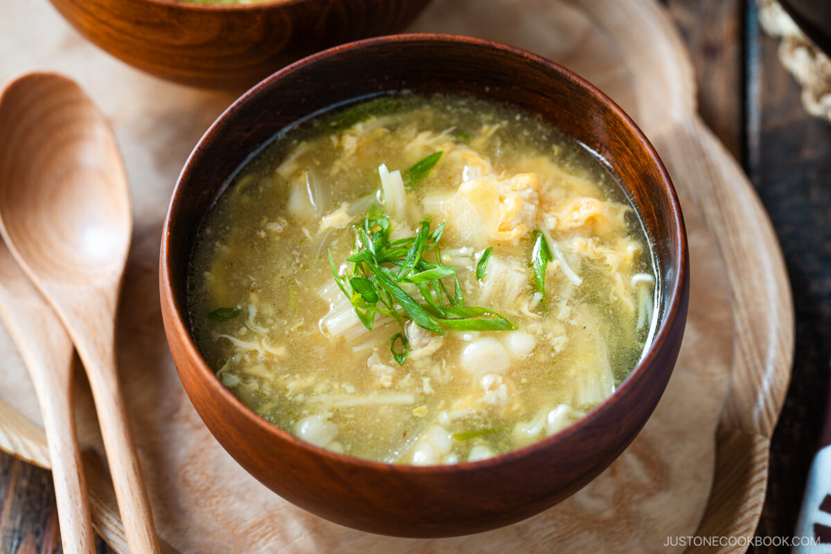 A wooden bowl filled with egg drop soup, garnished with chopped green onions. Two wooden spoons rest on a wooden tray beside the bowl.