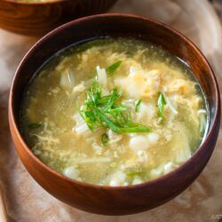 A wooden bowl filled with egg drop soup, garnished with chopped green onions. Two wooden spoons rest on a wooden tray beside the bowl.