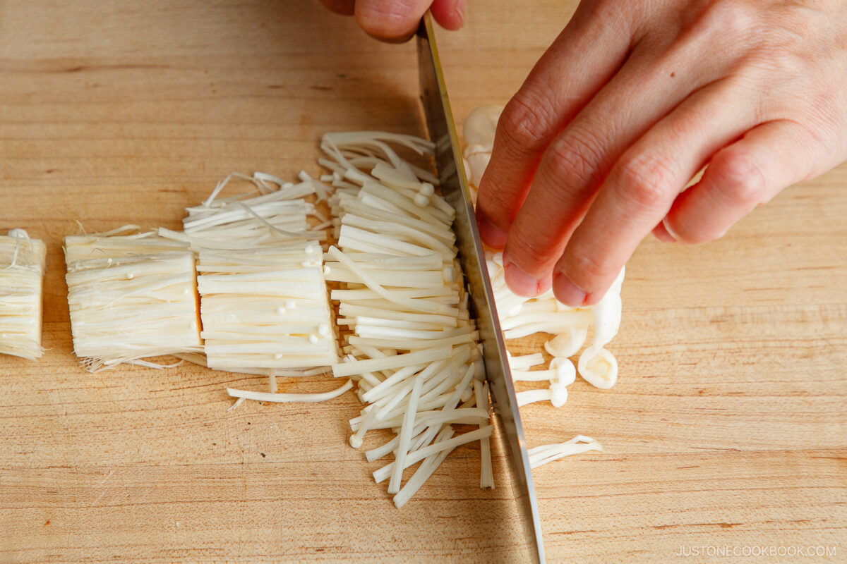 A person’s hands slicing enoki mushrooms on a wooden cutting board with a large knife.