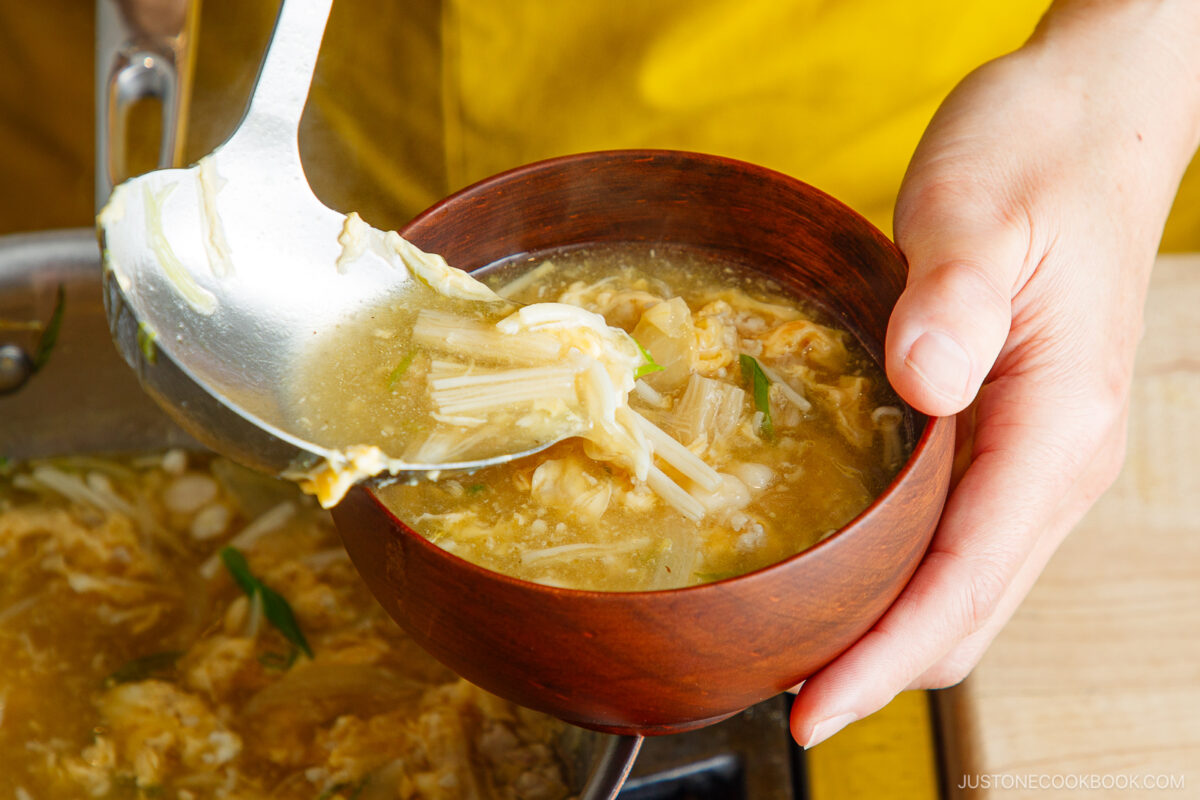 A hand holds a wooden bowl while soup with mushrooms, egg ribbons, and green onions is being ladled into it. The background shows a yellow fabric and part of the soup pot.