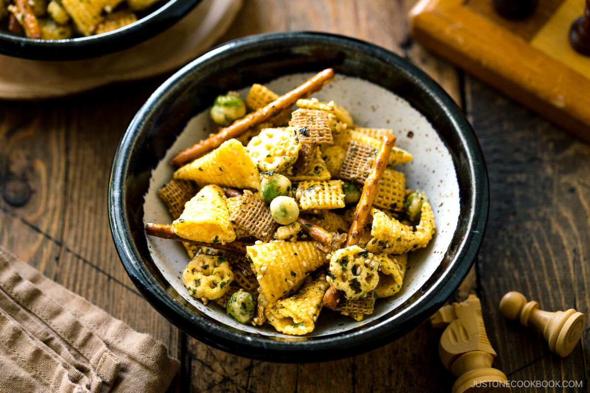A bowl of snack mix featuring Chex cereal, pretzel sticks, rice crackers, and green wasabi peas sits on a wooden table next to a napkin and a chessboard, with warm natural lighting.